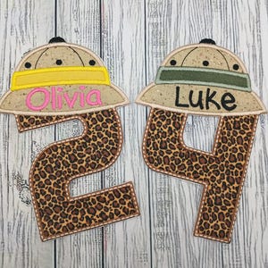 May include: Two embroidered safari-style hats with the names "Olivia" and "Luke" on them. The hats have a tan base with yellow and green bands. The numbers "2" and "4" are below the hats, with a leopard print pattern.