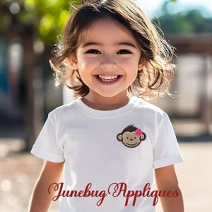 May include: A young girl wearing a white t-shirt with a brown monkey applique with a pink flower on its head. The text "Junebug Appliques" is visible at the bottom of the image.