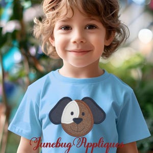 May include: A young child wearing a light blue t-shirt with an embroidered brown, white, and black dog applique. The dog has a brown nose and a friendly expression.