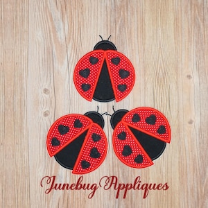 May include: Three red and black ladybug appliques with black heart details. The appliques are on a wood background.