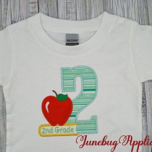 May include: White t-shirt with a green and white striped number "2" and a red apple with a green stem. Below the number is a yellow rectangle with the text "2nd Grade".