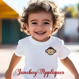 May include: A smiling child wearing a white t-shirt with a monkey face applique. The text "Junebug Appliques" is written in red script at the bottom of the shirt. The child is outside with a blurred background.