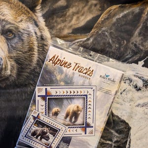 May include: A clear plastic package containing Alpine Tracks Border quilt pattern paper by Riley Blake Designs. The packaging features images of grizzly bears in a mountain landscape, with text detailing compatibility with 22 to 36 inch fabric panels.