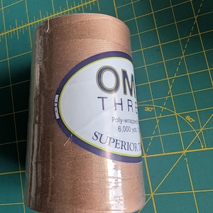 May include: A large spool of copper-colored sewing thread wrapped in clear plastic. The white label features the text OMNI THREAD, Poly-wrapped, 6,000 yds, and SUPERIOR THREADS, with Made in Vietnam printed vertically along the side of the oval label.