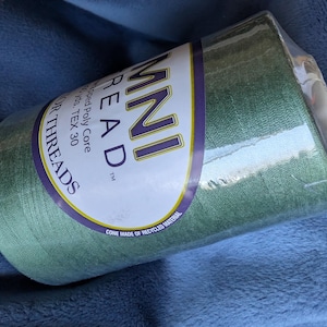 May include: A large cone of sage green Omni Thread wrapped in clear plastic. The white label features the text OMNI THREAD, Wrapped Poly Core, 6000 yds, TEX 30, and Cone Made of Recycled Material. The thread is a solid, muted green color.