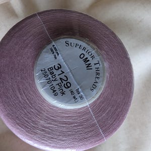 May include: A spool of dusty pink sewing thread labeled Superior Threads Omni 3129 Baby Pink, 6,000 yds, 100% Poly, Tex 30, 40 wt. The thread is wrapped tightly around a cylindrical core and is covered by a clear plastic film for protection.