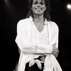 May include: Black and white image of a smiling person on stage, wearing a white shirt and black pants. The person is holding a microphone and has curly hair. The background is dark with stage lights.