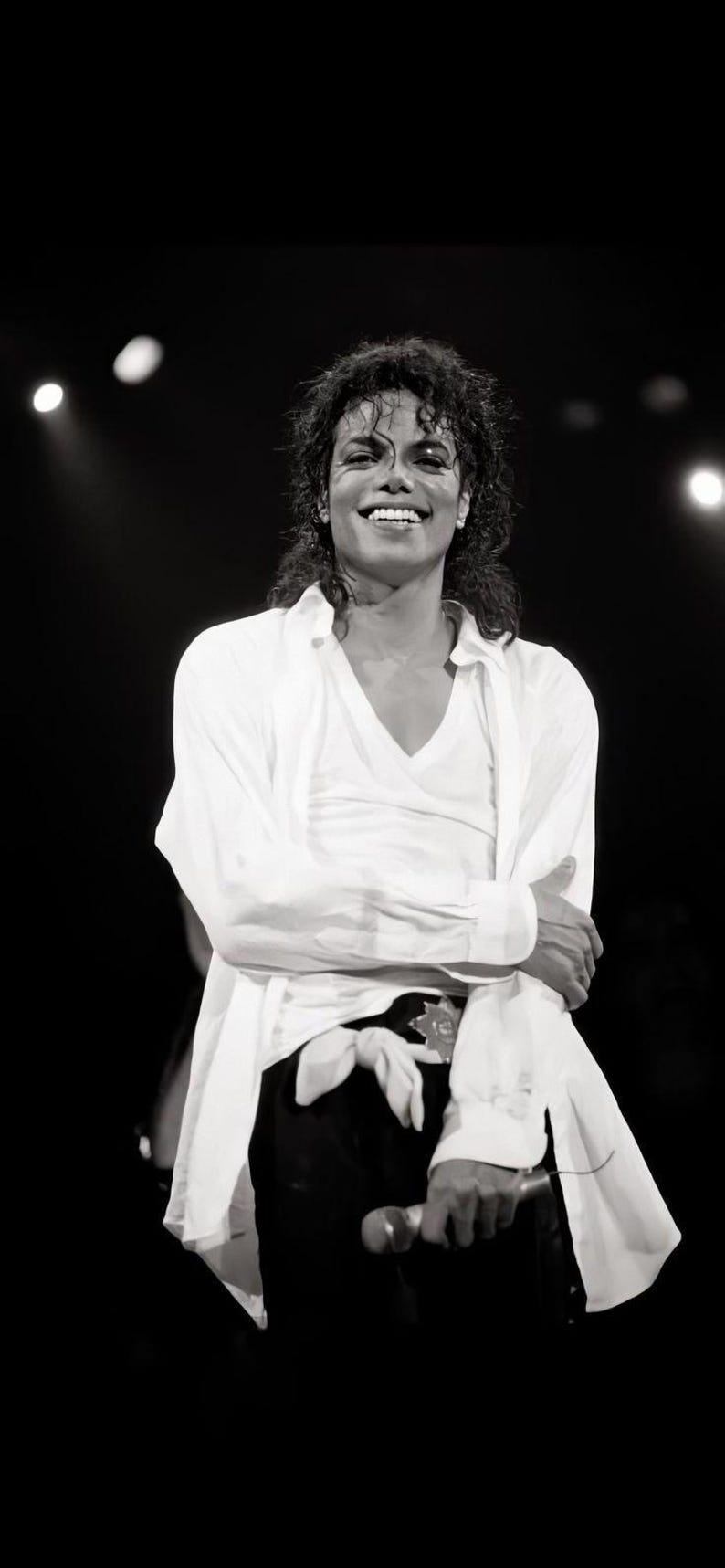 May include: Black and white image of a smiling person on stage, wearing a white shirt and black pants. The person is holding a microphone and has curly hair. The background is dark with stage lights.