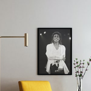 May include: Black and white framed photograph of a smiling person in a white shirt and black pants. The person is on a stage with lights in the background. A gold lamp and yellow chair are in the foreground.