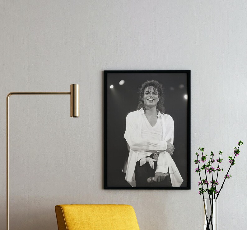 May include: Black and white framed photograph of a smiling person in a white shirt and black pants. The person is on a stage with lights in the background. A gold lamp and yellow chair are in the foreground.