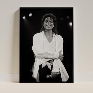May include: Black and white photograph of a smiling person wearing a white shirt and black pants, holding a microphone. The person is on a stage with spotlights in the background.