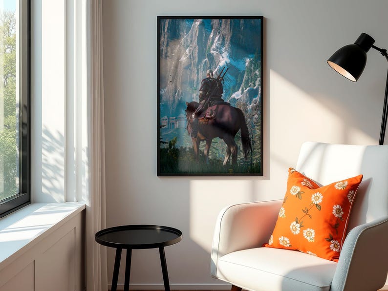 May include: A framed vertical wall art print featuring a digital illustration of a figure with long white hair riding a brown horse toward a snowy mountain range. The artwork is displayed in a black frame on a plain wall above a white armchair.
