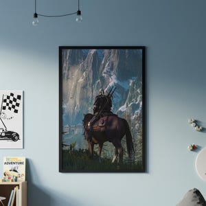 May include: A framed vertical wall art print depicting a figure with long white hair riding a brown horse, viewed from behind. The rider carries two swords on their back, facing tall, snow-capped mountain peaks and a stone fortress structure.
