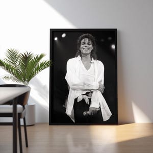 May include: Black and white framed photograph of a smiling person on stage, wearing a white shirt and black pants. The person is holding a microphone. The photograph is displayed on a white wall.