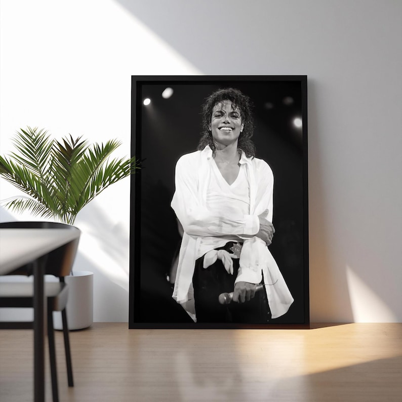 May include: Black and white framed photograph of a smiling person on stage, wearing a white shirt and black pants. The person is holding a microphone. The photograph is displayed on a white wall.
