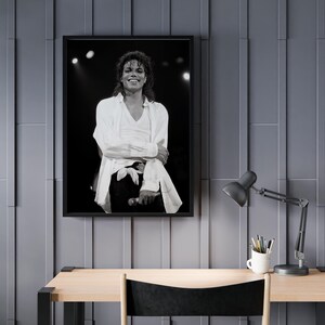 May include: Black and white framed photograph of a smiling person on stage, wearing a white shirt and black pants. The photograph is hanging on a gray wall above a wooden desk with a black lamp and a white mug.