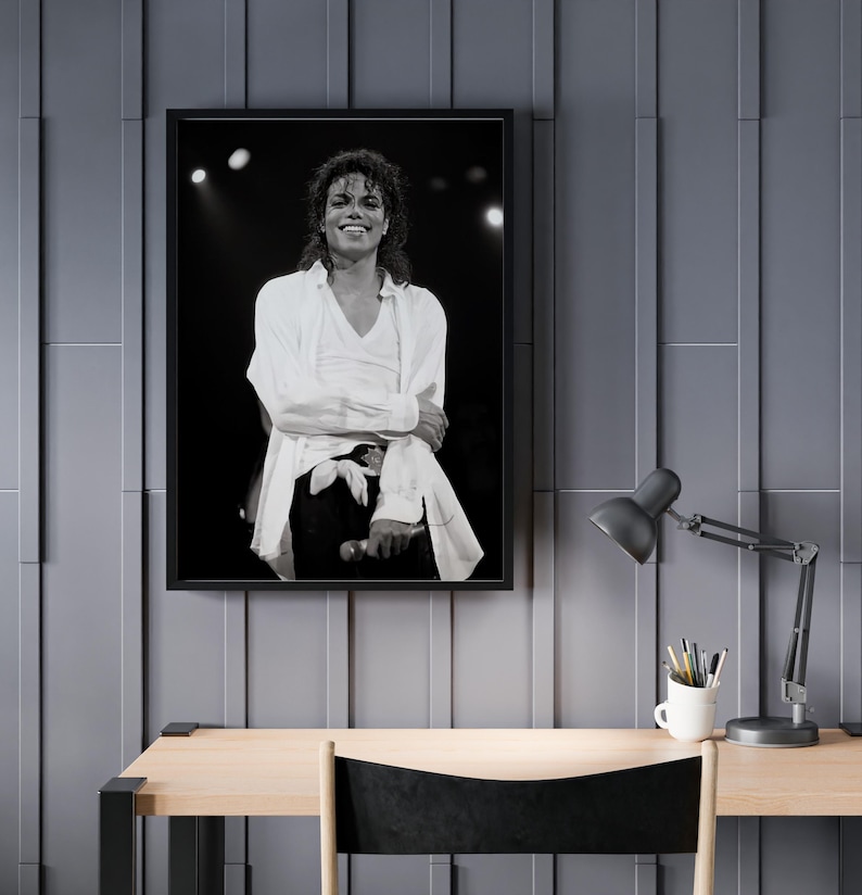 May include: Black and white framed photograph of a smiling person on stage, wearing a white shirt and black pants. The photograph is hanging on a gray wall above a wooden desk with a black lamp and a white mug.