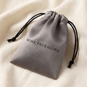 May include: A grey satin drawstring pouch with the text "WINK PACKAGING" printed on the front. The pouch has black drawstrings and is set against a cream-coloured fabric background. Suitable for jewellery or small gifts.