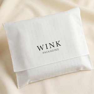 Puede incluir: Una bolsa de tela blanca con el texto "WINK PACKAGING" impreso en negro. La bolsa está doblada y el fondo es una tela beige claro.