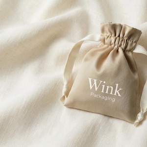 May include: A beige drawstring pouch with the text "Wink Packaging" printed in white. The pouch is made of a soft fabric and has a gathered top. The background is a textured, off-white fabric.