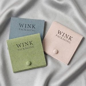 Pode incluir: Três bolsas quadradas de feltro nas cores azul acinzentado, verde sálvia e rosa suave. Cada bolsa possui um fecho de botão único e o texto WINK PACKAGING impresso em letras escuras na aba frontal destes pequenos recipientes de tecido.