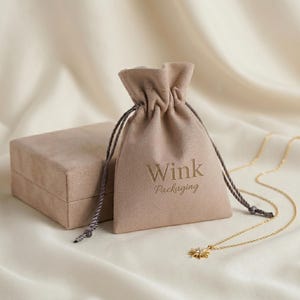 May include: A beige velvet drawstring pouch with the text Wink Packaging printed in gold, accompanied by a matching beige velvet square jewellery box and a delicate gold chain necklace featuring a small sunburst pendant with a central stone.