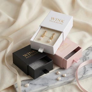 May include: Three jewelry boxes in white, black, and pink, each with the "WINK PACKAGING" logo in gold. The white box is open, displaying gold earrings with pearls. Several loose pearls are scattered on a marble surface.