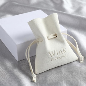 May include: A cream-coloured velvet drawstring pouch featuring the embroidered text Wink Packaging in a matching thread colour, positioned next to a plain white rectangular cardboard box, both designed for small item storage and presentation.