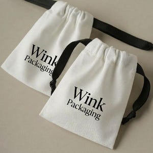 Puede incluir: Dos bolsas blancas con cordón y cordones negros. Las bolsas tienen las palabras "Wink Packaging" impresas en negro. Las bolsas están hechas de un material suave y texturizado.