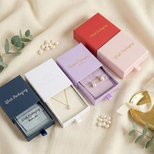 Peut inclure: Quatre boîtes à bijoux coulissantes carrées de couleur bleu marine, blanc, lavande et rose, portant chacune l'inscription dorée Wink Packaging. Les boîtes contiennent une bague en argent, un collier en or et des boucles d'oreilles en perles.