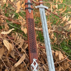 Può includere: Una spada con lama in acciaio Damasco a motivi e impugnatura rivestita in pelle marrone, accompagnata da un fodero in legno abbinato. Il fodero presenta intricati intagli a nodo celtico marrone scuro e finiture in metallo argentato.