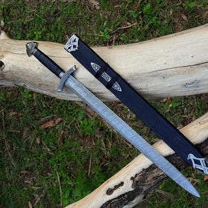 Può includere: Una spada color argento con impugnatura nera e lama in metallo decorata, accompagnata da un fodero nero con inserti in metallo argentato sulla punta e sull'imboccatura. La spada presenta una guardia e un pomolo con dettagli metallici.