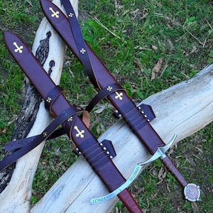 Peut inclure: Deux épées avec des fourreaux en cuir marron ornés d'emblèmes de croix dorées et de lanières en cuir. Les gardes en argent portent l'inscription "CHRISTUS VINCIT". Chaque épée possède une poignée gainée de cuir marron et un pommeau circulaire.