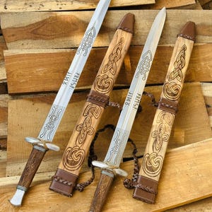 May include: Two silver-toned metal swords with pointed blades and brown textured handles. Each sword features etched knotwork patterns and runic text on the blade. They are accompanied by two matching wooden scabbards with brown leather accents.