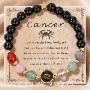 May include: A beaded bracelet featuring black spherical beads, a gold star charm, and a central circular pendant with the Cancer zodiac symbol. The bracelet includes colorful accent beads in red, pink, green, and clear, displayed on a card with text.