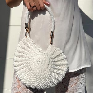 May include: A white, handmade crochet handbag with a distinctive fan-shaped design. The bag has a braided white handle with gold-coloured hardware. The bag is held up by a person.