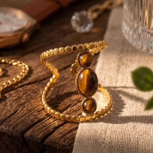 Puede incluir: Un brazalete abierto de tono dorado con una banda texturizada estilo cuentas y una pieza central vertical engastada con tres piedras preciosas de ojo de tigre marrón, lisas y ovaladas. La banda de metal tiene un patrón circular repetido.