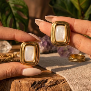 May include: A pair of rectangular gold-toned earrings featuring a smooth, iridescent white mother-of-pearl inlay centered within a polished, tiered metal frame. The earrings have a substantial, rounded border design with a warm, reflective finish.