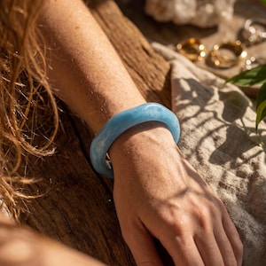 Puede incluir: Un brazalete rígido de color azul claro con un acabado marmolado semitranslúcido. El accesorio circular presenta un perfil grueso y redondeado con una superficie brillante que refleja la luz natural, creando sutiles variaciones en el tono azul.