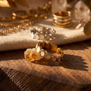Puede incluir: Anillos dorados y plateados con incrustaciones circulares blancas se exhiben sobre una superficie de madera. Un anillo dorado descansa sobre un cristal rugoso de color amarillo-naranja. Otras joyas, incluyendo una cadena de oro y anillos apilados, están en el fondo.