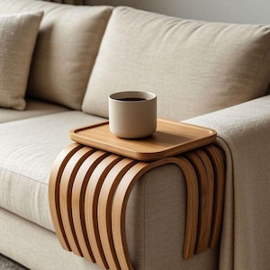 Puede incluir: Una mesa de brazo de sofá de bambú marrón claro con una bandeja a juego que sostiene una taza beige. La mesa tiene un diseño curvo y está colocada en el brazo de un sofá beige. La taza contiene un líquido oscuro.