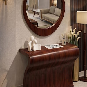 Può includere: Scena di ingresso moderna con un mobile consolle in legno scuro dal design curvo, sormontato da una scultura bianca e una stampa incorniciata. Un grande specchio rotondo è appeso sopra. Sono visibili anche una lampada dorata e una lampada da terra.