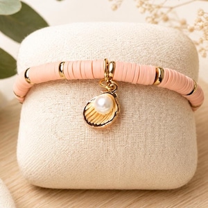 May include: A bracelet made of light pink disc-shaped beads interspersed with small gold-toned metal spacers. A gold-toned metal charm shaped like a seashell with a single white pearl nestled inside hangs from the centre of the beaded bracelet.