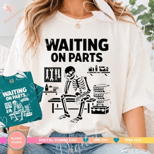 May include: Cream-colored t-shirt with the text "WAITING ON PARTS" in black. The graphic features a skeleton sitting on a bench, surrounded by tools, tires, and a toolbox. A smaller teal shirt with the same design is visible.