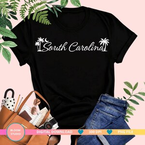 Op de afbeelding: Een zwart T-shirt met korte mouwen en witte tekst South Carolina, geflankeerd door twee silhouetten van palmbomen en een maansikkel. Het ontwerp is gecentreerd op de borst van het T-shirt, dat plat op een lichte achtergrond ligt.
