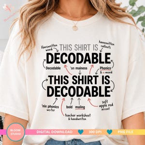 Op de afbeelding: Een crèmekleurig t-shirt met de zwarte tekst THIS SHIRT IS DECODABLE die twee keer wordt herhaald. Het ontwerp bevat diverse handgeschreven toelichtingen met pijlen die naar de tekst wijzen, gelabeld met termen zoals phonics, bold en maling.