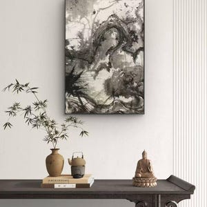 May include: A framed abstract ink wash painting in black and white hangs above a dark wooden console table. On the table sits a tan ceramic vase with bamboo branches, a small teapot on a stand, a stack of two books, and a bronze-colored Buddha statue.