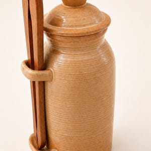 Puede incluir: Un tarro de cerámica de color beige con tapa y unas pinzas de madera. El tarro tiene forma cilíndrica con una superficie texturizada. Las pinzas se insertan en un lazo en el lateral del tarro. El tarro mide unos 20 cm de alto.