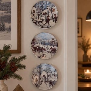 Könnte beinhalten: Drei kreisförmige Zierteller, die vertikal an einer Wand montiert sind und jeweils eine detaillierte Winterszene mit verschneiten Landschaften, Kiefern, Rentieren und Figuren in traditioneller Kleidung in Blau-, Weiß- und Brauntönen zeigen.