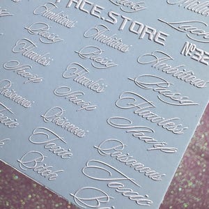 Op de afbeelding: Een vel zilveren nail art stickers met diverse cursieve woorden zoals "Lucky", "Ambitious" en "Toxic Bitch". De tekst "COOLSPACE.STORE" is ook zichtbaar. De stickers staan op een lichtblauwe achtergrond.